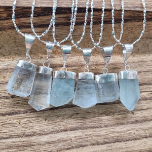 May include: Seven silver necklaces with aquamarine crystal pendants. The pendants are faceted and have a light blue color. The necklaces are on a wooden surface.