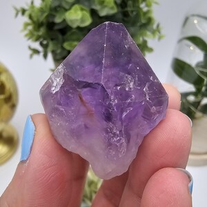 Pode incluir: Um cristal de ametista roxo com uma ponta pontiaguda e bordas ásperas. O cristal é uma formação natural de cor roxa intensa.