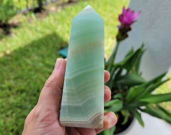 May include: A green and white banded calcite crystal point, also known as a tower, is held in a hand. The crystal is polished and has a smooth, glassy surface.