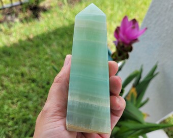 May include: A green and white banded calcite crystal point, also known as Mexican onyx, in the shape of a pyramid. The crystal is being held in a hand.