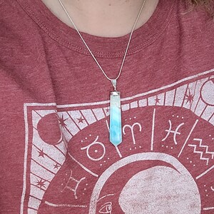 May include: A silver necklace with a pendant featuring a blue and white crystal. The crystal is rectangular with a pointed bottom. The person is wearing a maroon t-shirt with a white graphic of a moon and zodiac symbols.