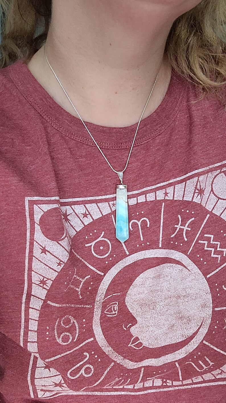 May include: A silver necklace with a pendant featuring a blue and white crystal. The crystal is rectangular with a pointed bottom. The person is wearing a maroon t-shirt with a white graphic of a moon and zodiac symbols.
