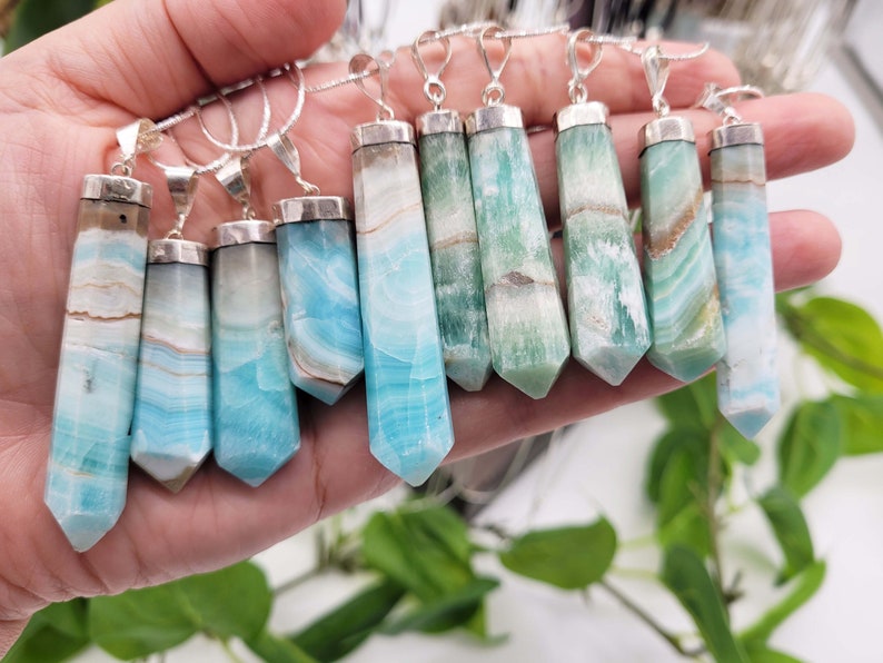 May include: A collection of gemstone pendants with silver-colored metal caps and bails. The pendants are rectangular with a pointed bottom and feature shades of blue, green, and brown. They are displayed against a blurred background.