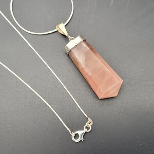 May include: A silver chain necklace with a pink crystal pendant. The pendant is a long, rectangular shape with a pointed end.