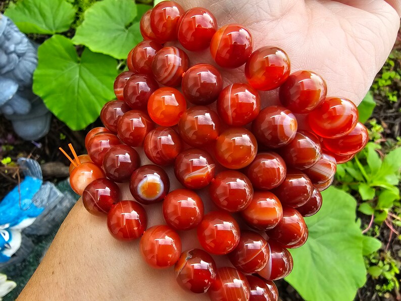 May include: A bracelet made of carnelian beads. The beads are a deep orange color with white stripes. The bracelet is on a person's wrist.