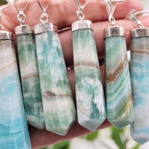 May include: A collection of gemstone pendants with silver-colored caps and bails. The pendants are rectangular with a faceted, tapered shape. The gemstones display a mix of blue, green, white, and brown colors, with a banded pattern. The pendants are on silver chains.