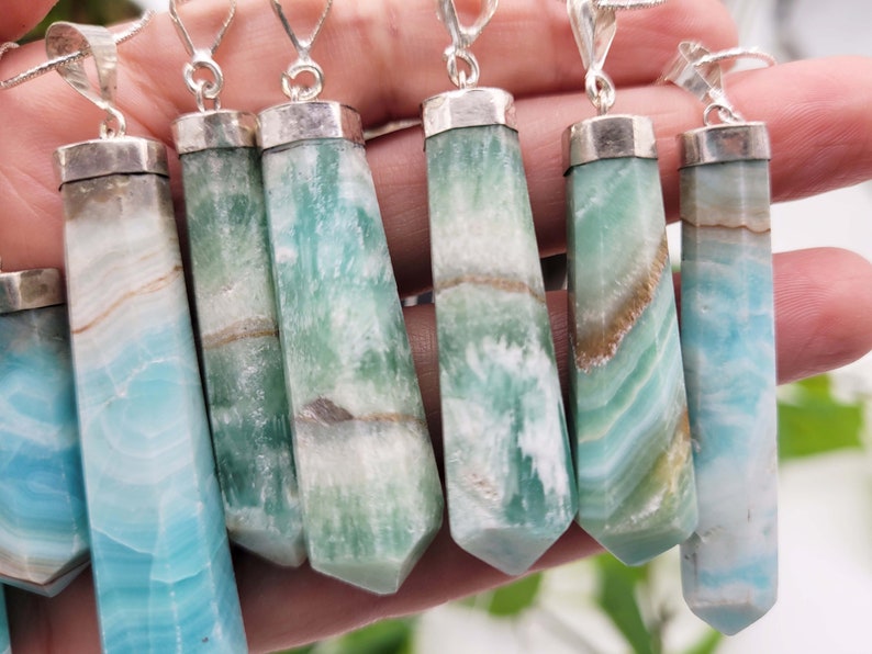 May include: A collection of gemstone pendants with silver-colored caps and bails. The pendants are rectangular with a faceted, tapered shape. The gemstones display a mix of blue, green, white, and brown colors, with a banded pattern. The pendants are on silver chains.