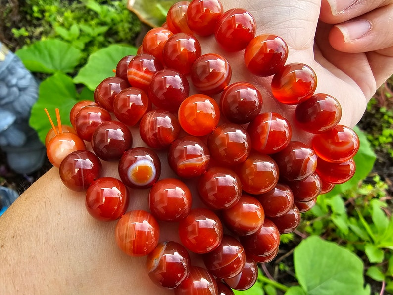May include: A strand of carnelian beads, featuring a mix of red and orange tones with white banding. The beads are round and smooth, with a polished finish.