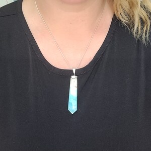 May include: A silver necklace with a pendant. The pendant is a light blue and white crystal in a hexagonal shape. The necklace is worn on a black top.