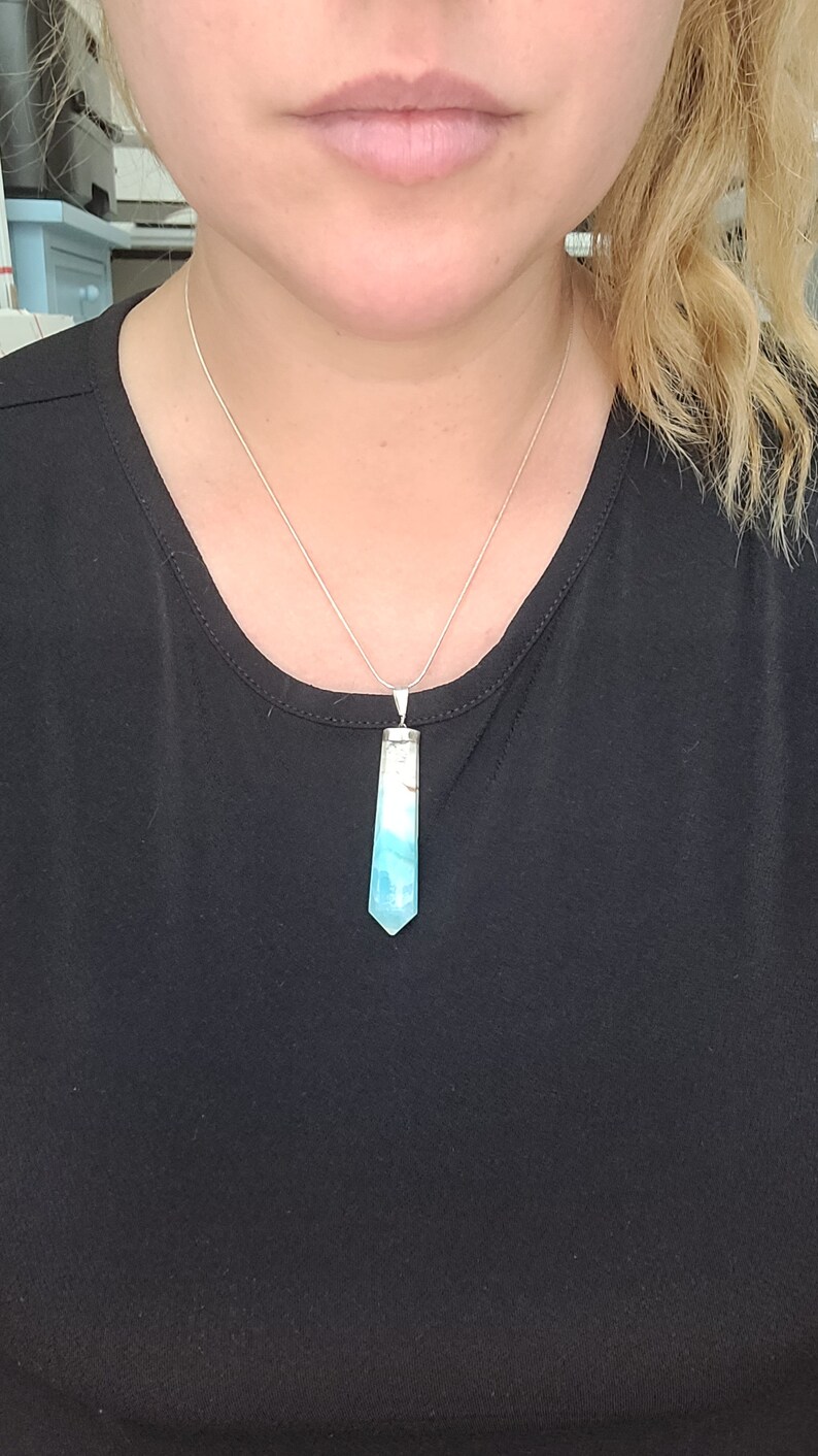 May include: A silver necklace with a pendant. The pendant is a light blue and white crystal in a hexagonal shape. The necklace is worn on a black top.