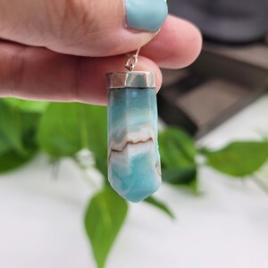 May include: A blue and white gemstone pendant with a silver cap. The gemstone has a unique pattern of light blue and brown swirls. The pendant is suspended from a silver hook, showcasing a natural crystal formation.