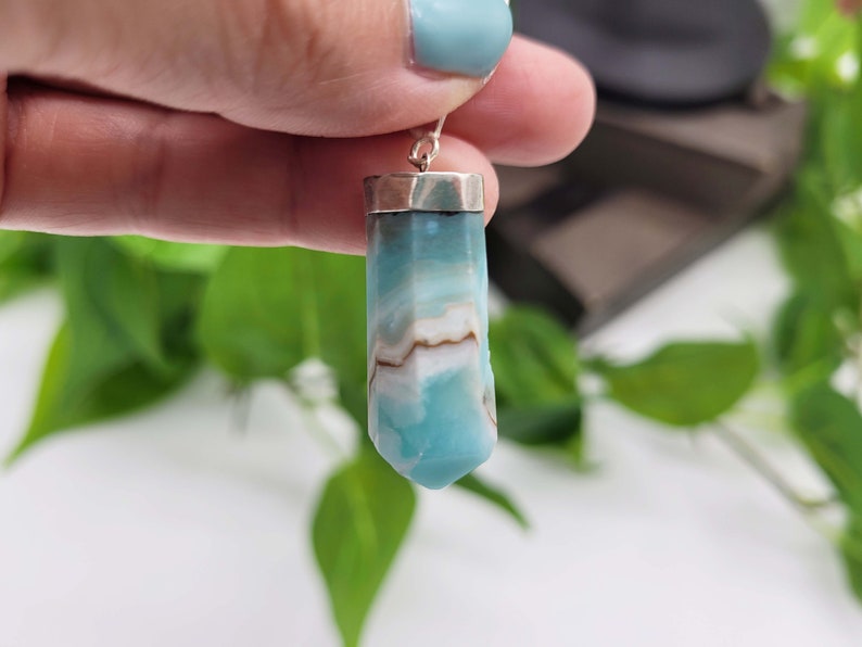 May include: A blue and white gemstone pendant with a silver cap. The gemstone has a unique pattern of light blue and brown swirls. The pendant is suspended from a silver hook, showcasing a natural crystal formation.