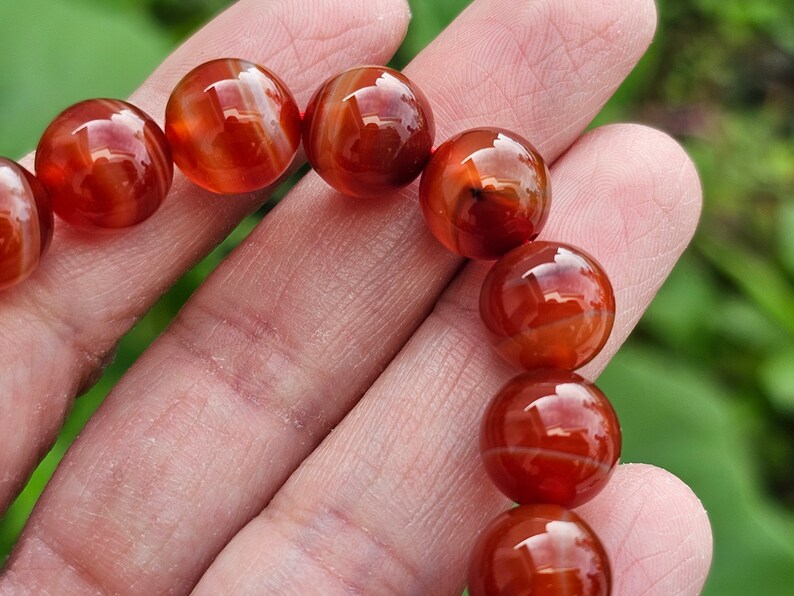 May include: A close-up of a red carnelian bracelet with round beads. The beads are polished and have a natural, striated pattern.