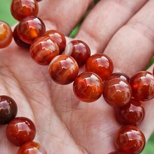May include: A strand of carnelian beads, with a reddish-orange color and white banding, is displayed on a hand. The beads are round and smooth.