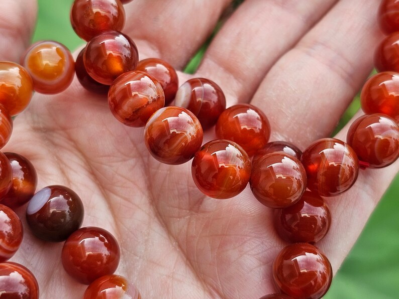 May include: A strand of carnelian beads, with a reddish-orange color and white banding, is displayed on a hand. The beads are round and smooth.