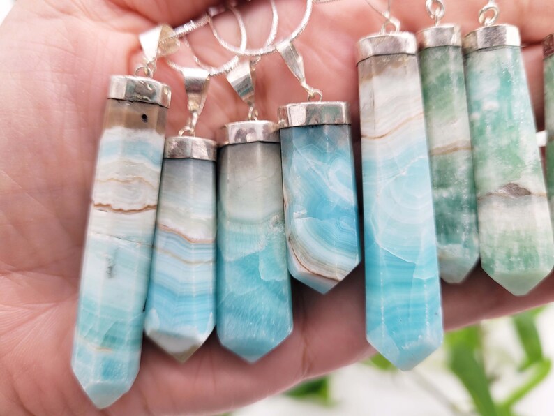May include: A collection of gemstone pendants with silver-colored metal caps and bails. The pendants are rectangular with a pointed bottom and feature bands of blue, white, and brown colors. Some pendants are green.