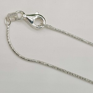 May include: A close-up of a silver necklace chain with a lobster clasp. The chain has a textured, snake-like design. The clasp is open, showing the details of the metal. The background is a plain, light color.
