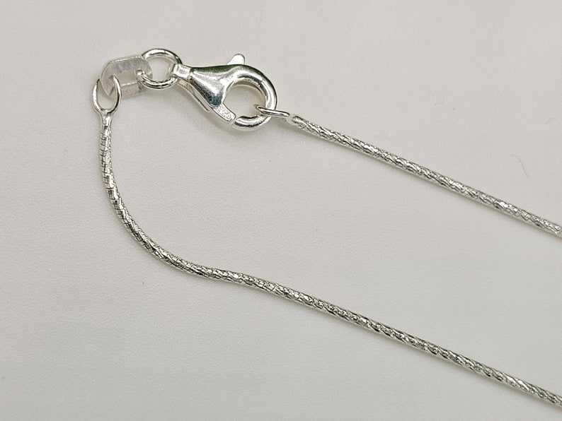 May include: A close-up of a silver necklace chain with a lobster clasp. The chain has a textured, snake-like design. The clasp is open, showing the details of the metal. The background is a plain, light color.