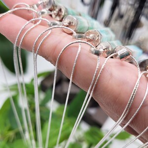 May include: Silver-toned necklaces with delicate chains and cylindrical pendants. The necklaces are held in a hand, with the chains cascading downwards. The pendants have a silver cap and a light green stone. The background is blurred with green foliage.