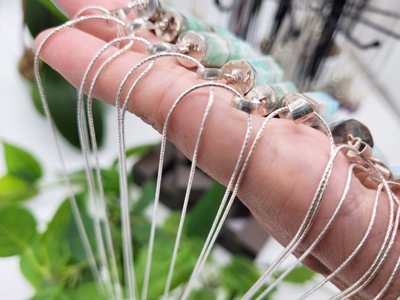 May include: Silver-toned necklaces with delicate chains and cylindrical pendants. The necklaces are held in a hand, with the chains cascading downwards. The pendants have a silver cap and a light green stone. The background is blurred with green foliage.