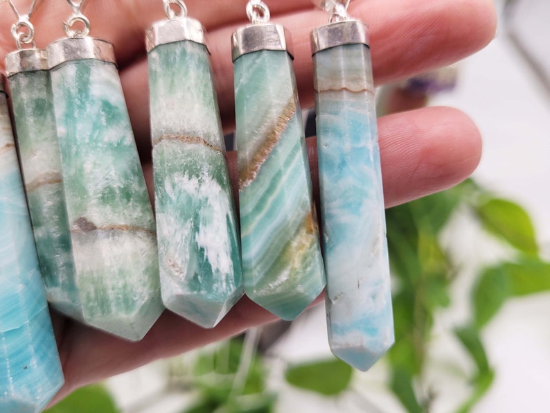 May include: A collection of gemstone pendants with silver-colored caps and chains. The pendants are hexagonal, with a variety of colors including light blue, green, and brown. The gemstones have a polished, smooth surface.