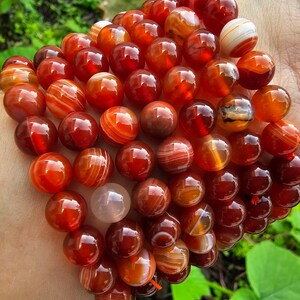May include: A bracelet made of carnelian beads. The beads are a deep red color with white stripes. The bracelet is stacked on a person's wrist.