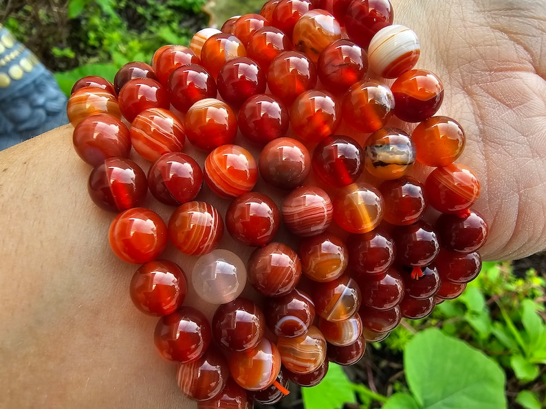 May include: A bracelet made of carnelian beads. The beads are a deep red color with white stripes. The bracelet is stacked on a person's wrist.