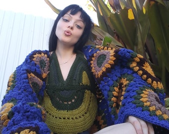 May include: A woman wearing a blue, green, brown, and yellow crocheted cardigan with a matching crocheted top. The cardigan has a granny square pattern.