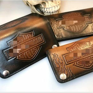 Può includere: Tre portafogli in pelle di varie dimensioni e tonalità di marrone. Ogni portafoglio presenta un logo in rilievo con le parole "MOTOR" e "CYCLES". I portafogli hanno un tema motociclistico e sono assicurati con chiusure a scatto argentate.