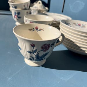 Puede incluir: Un juego de tazas de té y platillos blancos con diseños florales azules y rosas. Las tazas y los platillos están apilados sobre una superficie azul.