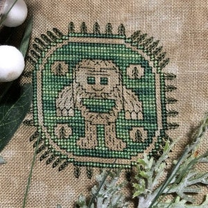 Peut inclure: Broderie au point de croix représentant une créature verte et brune avec une barbe et un gros nez, entourée d'un cercle vert avec une bordure brune. La créature se tient dans un décor de forêt avec des arbres et des buissons.