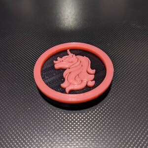 May include: A red circular button with a black unicorn silhouette in the center. The button is made of plastic and has a smooth, glossy finish.