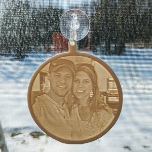 May include: A round, cream-colored ornament with a photo of a couple. The ornament is hanging from a suction cup on a window.