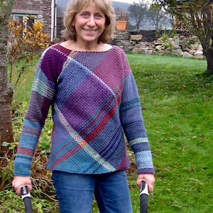 Puede incluir: Una mujer lleva un suéter multicolor con un patrón de cuadros en tonos de morado, azul, rojo y blanco. El suéter tiene un cuello redondo y mangas largas. Lleva pantalones vaqueros azules y sostiene dos bastones de senderismo.