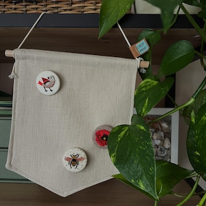 Könnte beinhalten: Ein beigefarbener Leinenbanner mit drei gestickten Applikationen: einem rotbrüstigen Vogel, einer roten Mohnblume und einer Biene. Der Banner hängt an einer Holzleiste und hat ein kleines braunes Etikett mit dem Text "EASY ideas" darauf.