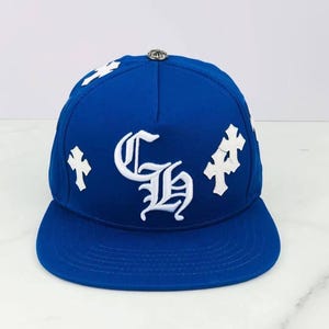 May include: A royal blue baseball cap featuring white embroidered gothic-style lettering of the initials CH in the center. The cap is decorated with white embroidered cross symbols on the front panels and has a silver-toned metal button on top.