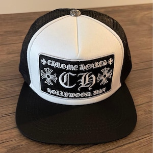 Peut inclure: Une casquette trucker noire et blanche avec un panneau avant blanc et un écusson rectangulaire noir. L'écusson affiche le texte CHROME HEARTS, les lettres CH et HOLLYWOOD, USA en broderie blanche, surmonté d'un bouton en métal argenté.