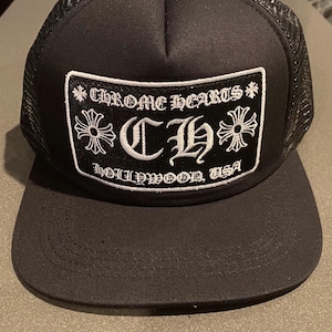 May include: A black trucker hat featuring a mesh back and a solid front panel. A rectangular patch is embroidered with the white text CHROME HEARTS, the letters CH, and the text HOLLYWOOD, USA, flanked by two cross symbols and a silver button on top.