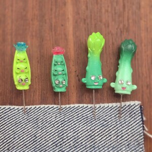 May include: Four green and white plastic pins with cartoon faces of peas and asparagus. The pins are on a wooden surface and a blue denim fabric.