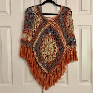May include: A crocheted poncho with a geometric pattern in shades of orange, brown, beige, and blue. The poncho has fringe around the edges.