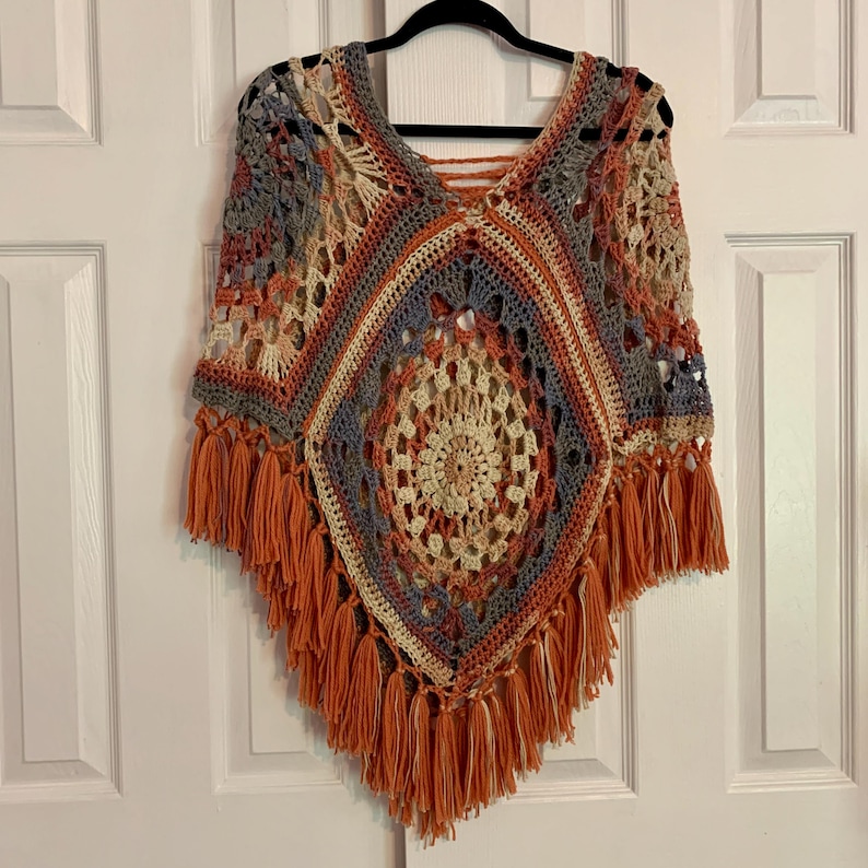 May include: A crocheted poncho with a geometric pattern in shades of orange, brown, beige, and blue. The poncho has fringe around the edges.