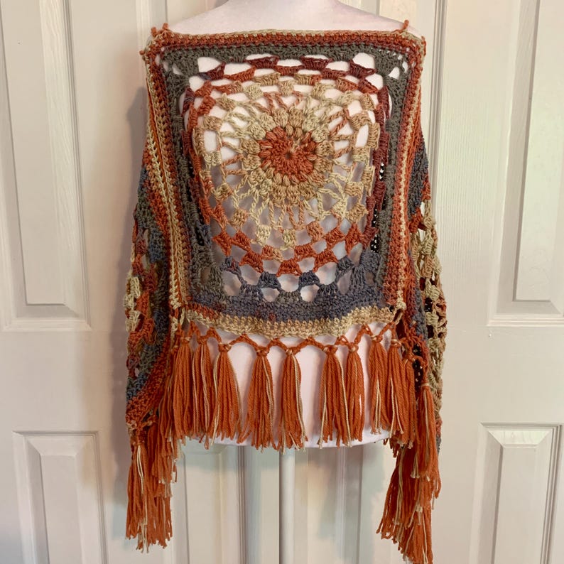 May include: A crocheted poncho with a circular design in shades of orange, brown, beige, and gray. The poncho has a fringe border.