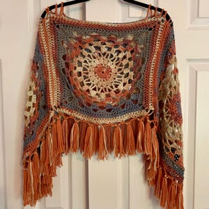 May include: A crocheted poncho with a circular pattern in shades of orange, brown, beige, and blue. The poncho has a fringe border in orange and beige.