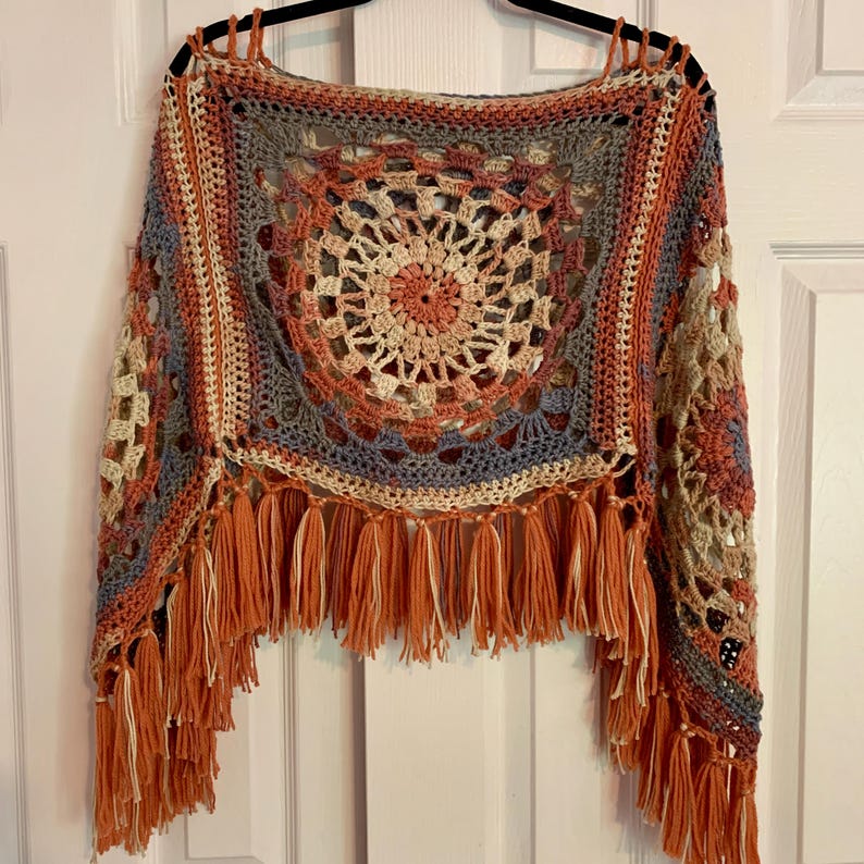 May include: A crocheted poncho with a circular pattern in shades of orange, brown, beige, and blue. The poncho has a fringe border in orange and beige.