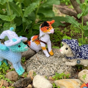May include: Three crocheted fantasy creatures: a blue dragon wearing a wizard hat, a calico cat with a saddle, and a white mouse in a blue hooded cloak. The creatures are displayed on rocks with greenery.