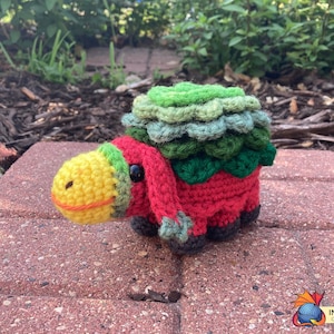 Puede incluir: Un animal de peluche tejido a crochet con un cuerpo rojo, una cabeza verde y amarilla y una espalda con forma de caparazón verde y marrón. El animal está sentado sobre una superficie de ladrillo.