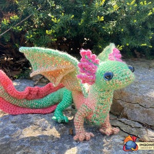 May include: A crocheted dragon figurine in shades of green, pink, and orange. The dragon has large, detailed wings, a long tail, and small feet. The dragon is posed on a rock, with a natural background.