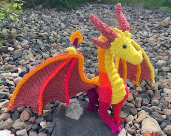 Könnte beinhalten: Ein gehäkeltes Drachenspielzeug aus gelbem, orangem und rotem Garn. Der Drache hat Flügel und einen langen Schwanz. Der Drache sitzt auf einem Bett aus kleinen Steinen.