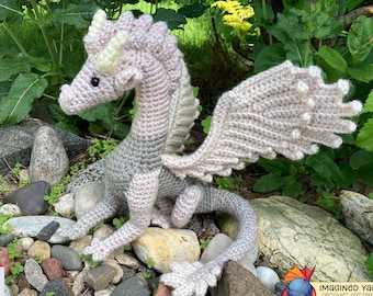 May include: A crocheted dragon amigurumi toy with a light pink and gray color gradient. The dragon is sitting on a bed of small rocks with its wings spread out. The dragon has a long tail and a small, pointed snout. The toy is made with a soft yarn and has a cute, whimsical expression.