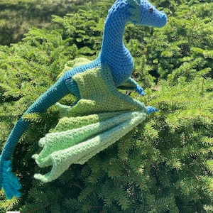 Peut inclure: Un dragon bleu et vert en crochet avec les ailes d&eacute;ploy&eacute;es. Le dragon est assis sur un lit de branches de pin vertes.